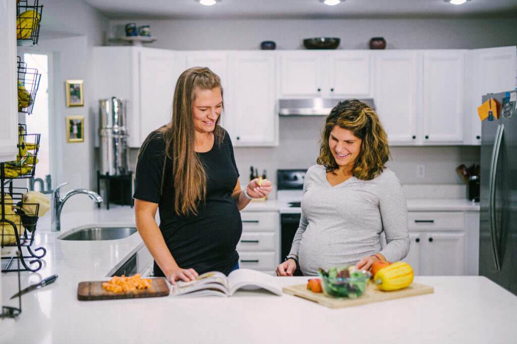 Two pregnant women looking at a cookbook at the kitchen counter.